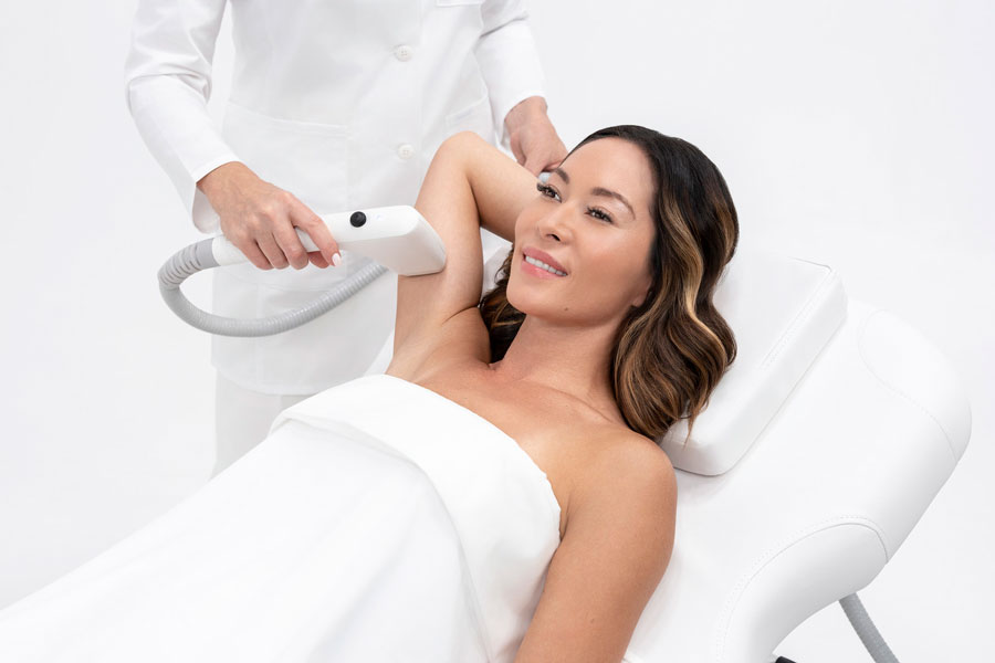 Woman lies on a white treatment chair while a clinician uses a handheld device on her armpit for a cosmetic procedure/laser treatment.