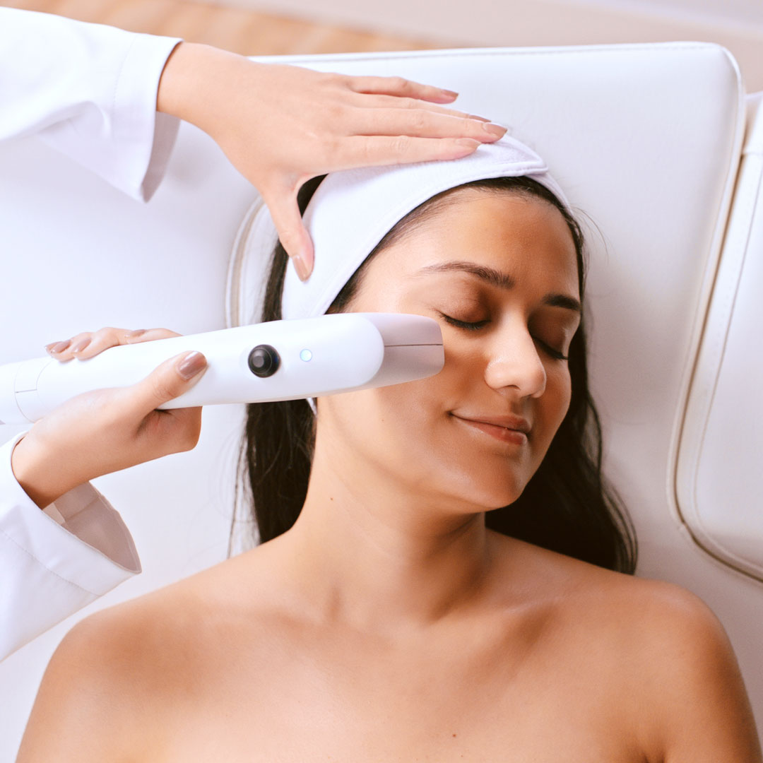 Woman lying with eyes closed as a clinician uses a handheld device on her cheek during a facial treatment.