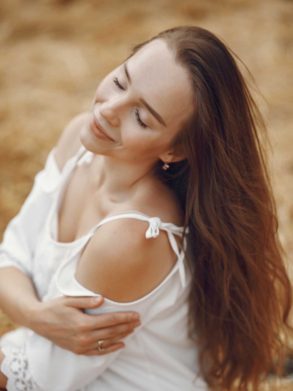 Portrait of a smiling woman with long brown hair, wearing a white top, outdoors in warm sunlight with eyes closed